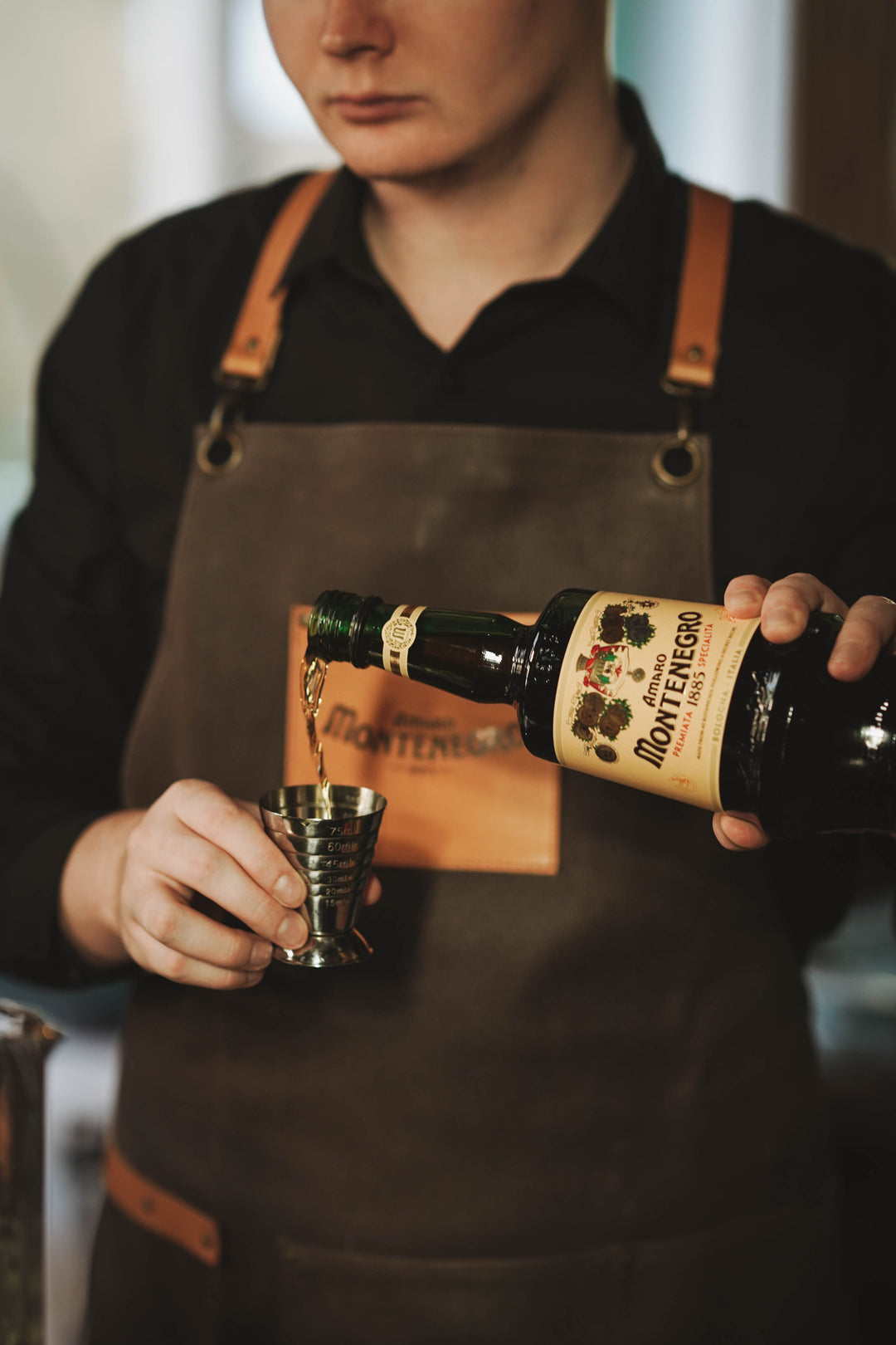 Person in a brown apron pouring a drink from a bottle labeled 'Montenegro Aperitivo' into a small cup.