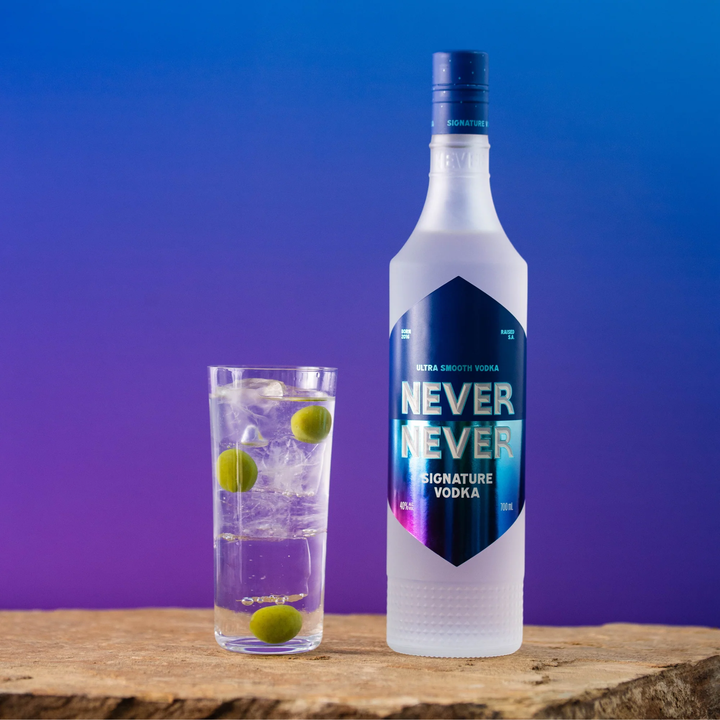 Bottle of Never Never Vodka with a glass of ice and green olives on a wooden surface against a gradient purple and blue background.